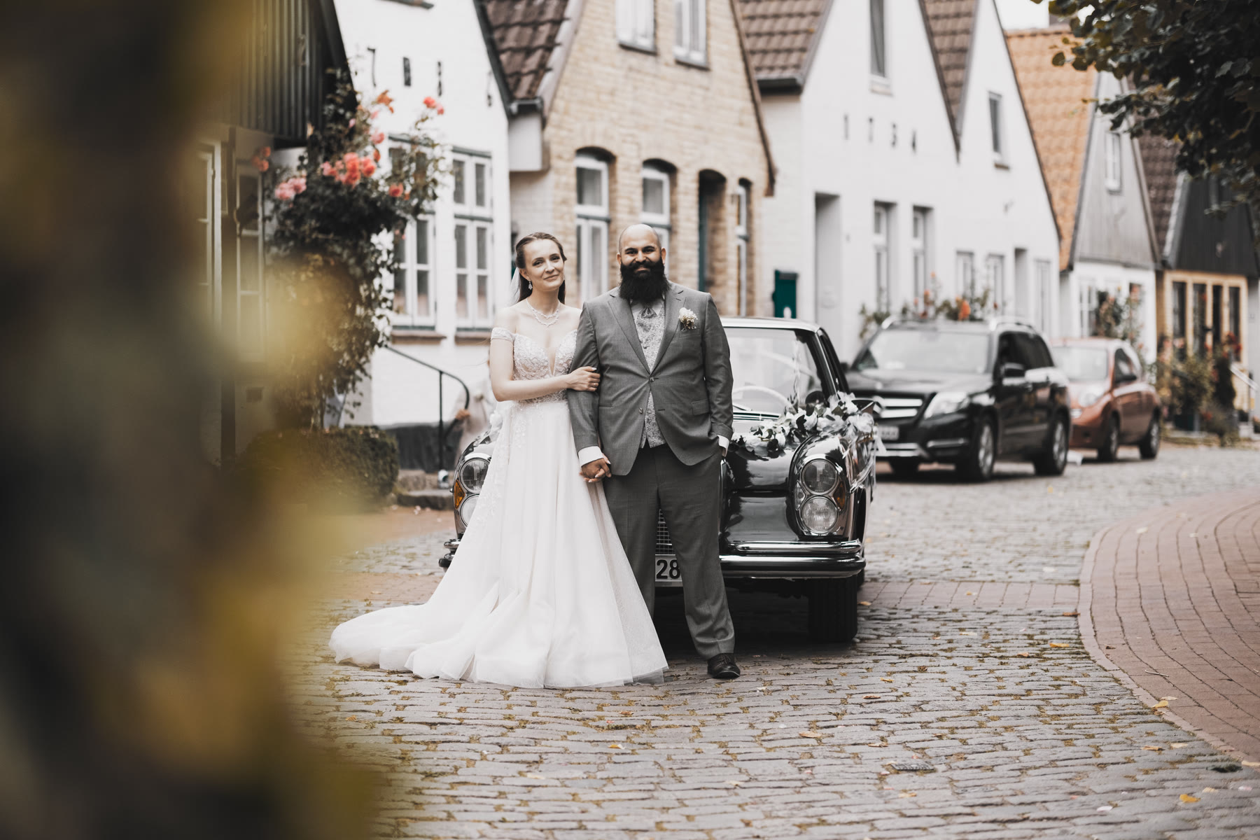 Brautpaar steht vor historischen Häusern in einer Altstadtgasse neben einem Hochzeits-Oldtimer