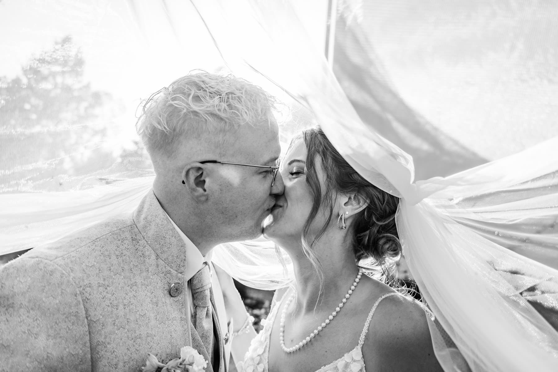 Brautpaar küsst sich unter einem wehenden Hochzeitsschleier im Gegenlicht, Schwarz-Weiß