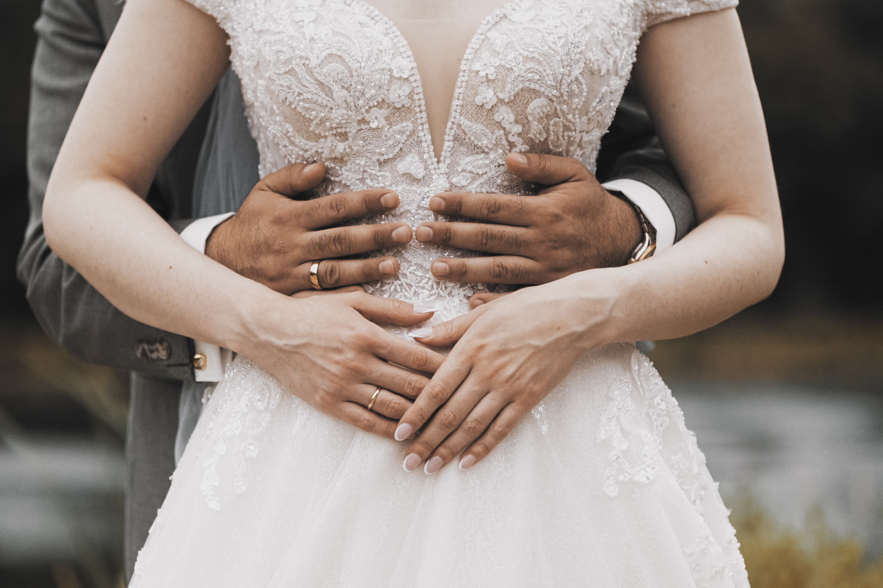 Detailaufnahme: ineinander verschränkte Hände des Brautpaars vor dem Hochzeitskleid