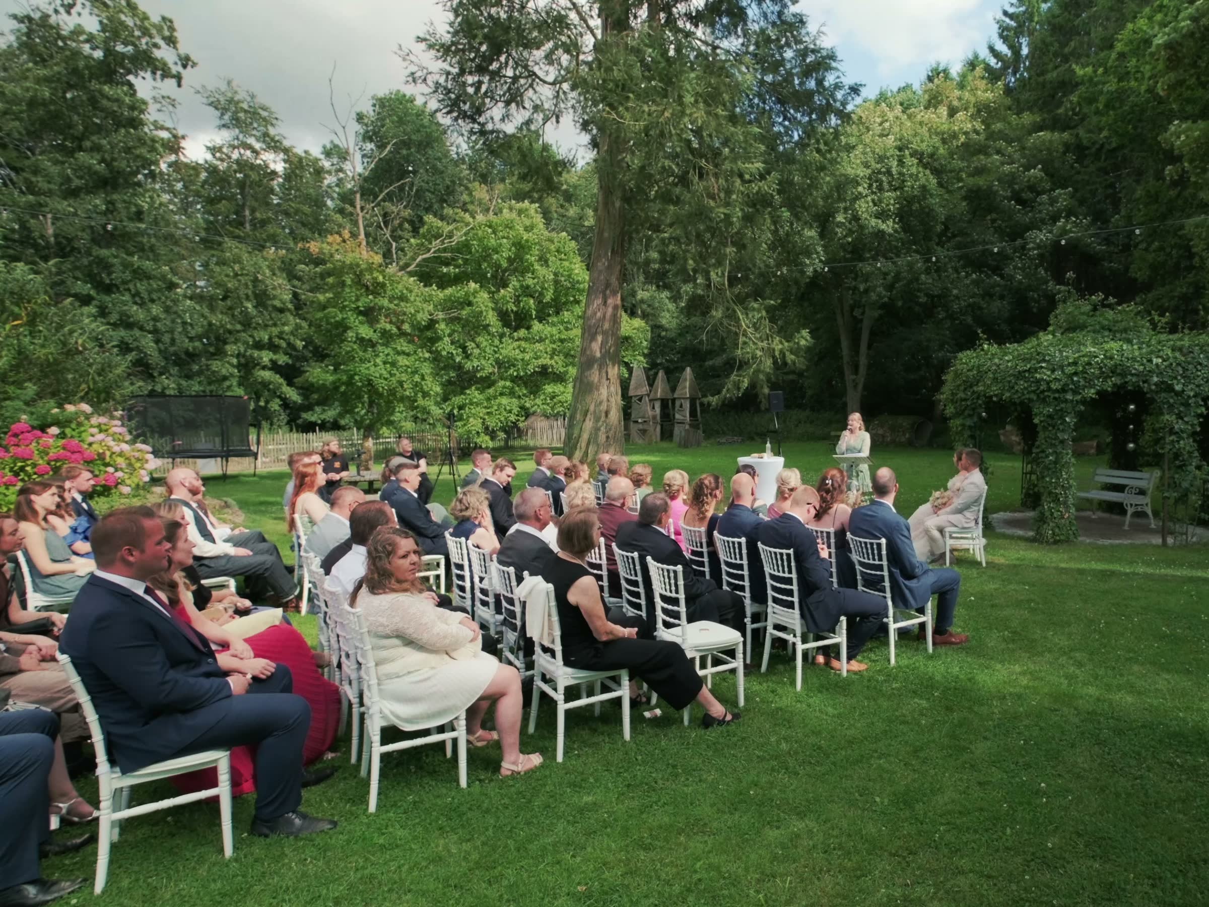 Standbild aus einem Hochzeitsfilm: freie Trauung im Garten mit Gästen auf weißen Stühlen